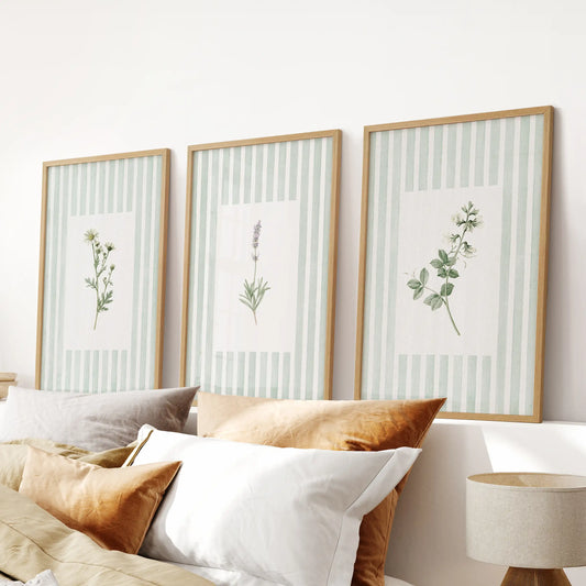 A set of three green floral botanical wall art prints in natural oak frames hangs over a bedroom headboard.