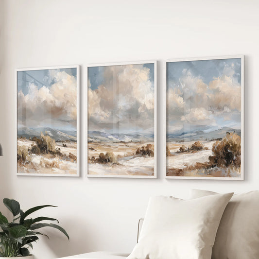 Rustic farmhouse scenery, a set of three prints in white frames, hangs over the couch to warm the living room with soft beige, brown, and blue tones.