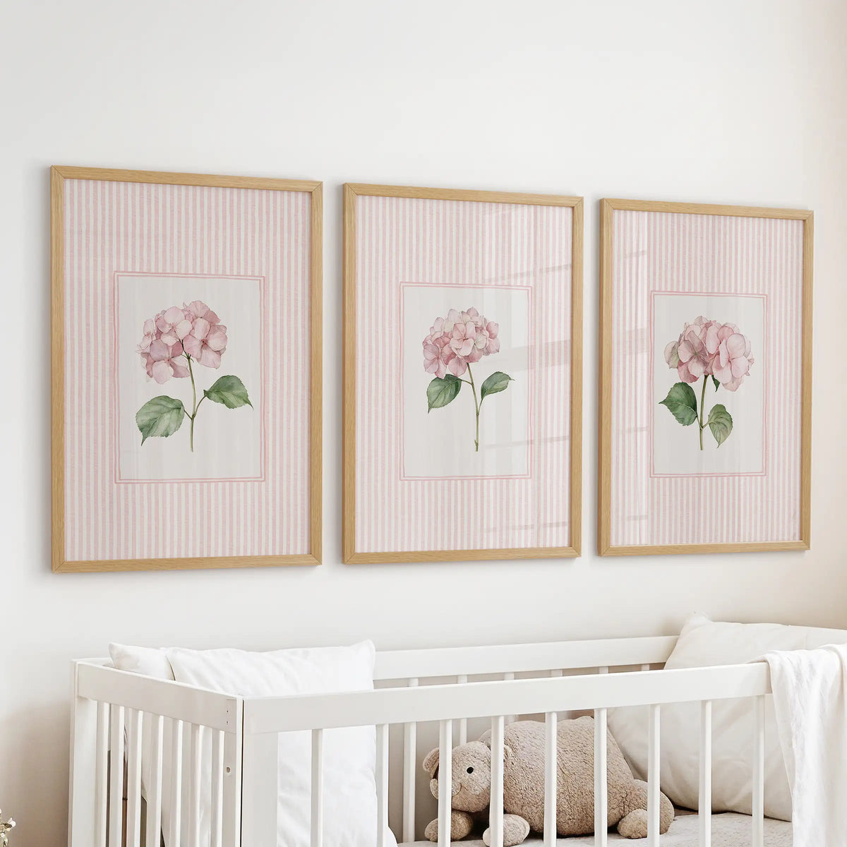 Three-piece wall art set of pink striped florals in light oak frames above a cot in an infant room.