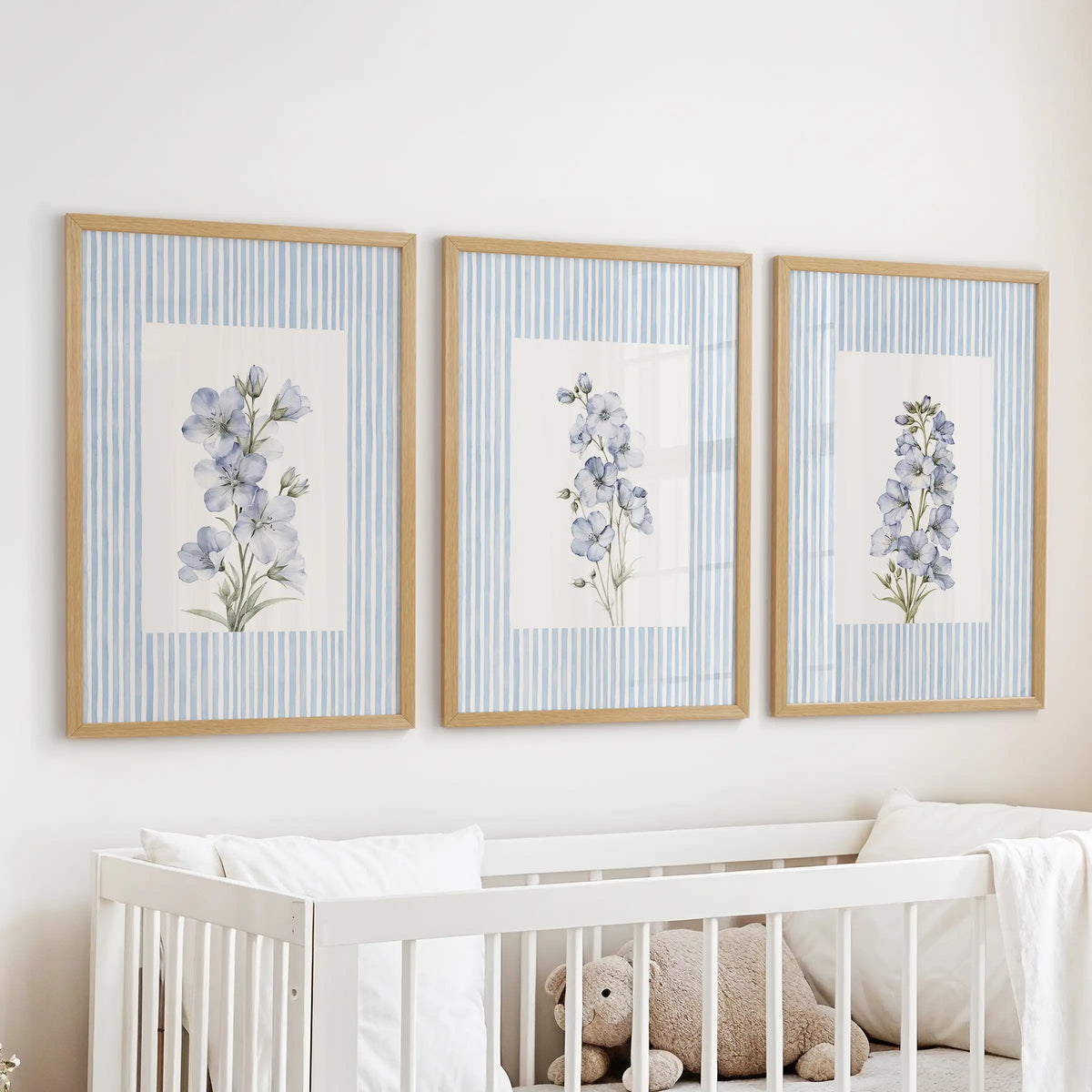 Set of three blue floral watercolor artworks in light oak frames, hanging above a white cot in a pale blue nursery.