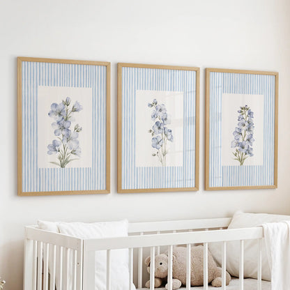 Set of three blue floral watercolor artworks in light oak frames, hanging above a white cot in a pale blue nursery.