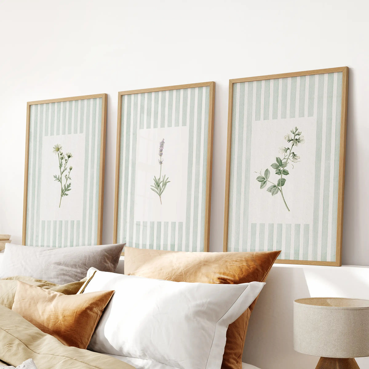 A set of three green floral botanical wall art prints in natural oak frames hangs over a bedroom headboard.