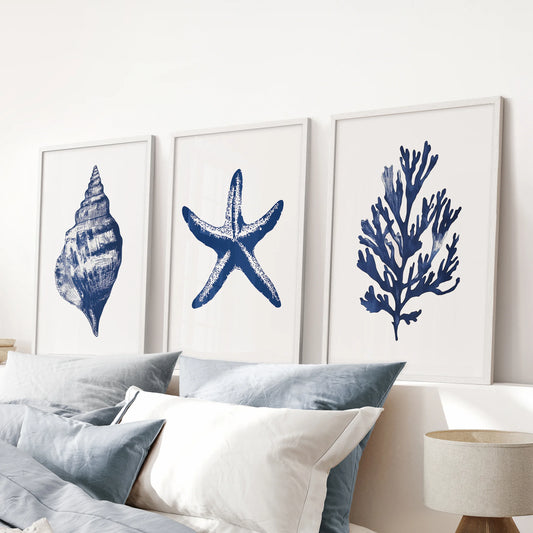 Navy blue beach house art three-piece print set comes as white framed prints for a blue bedroom, displayed over the bed headboard.