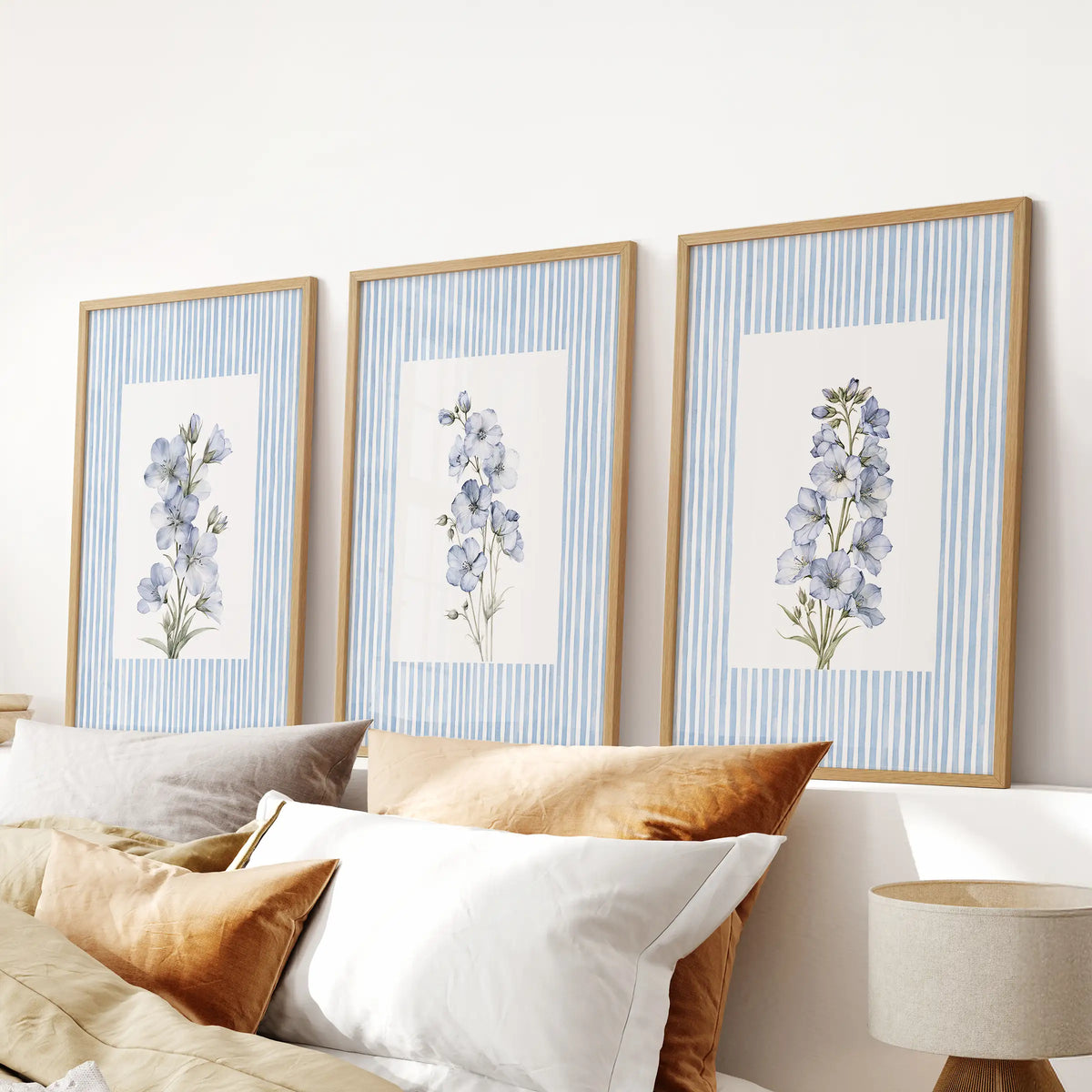 Three-piece pastel botanical wall art set in light oak frames, arranged above the bed in a pale blue bedroom.