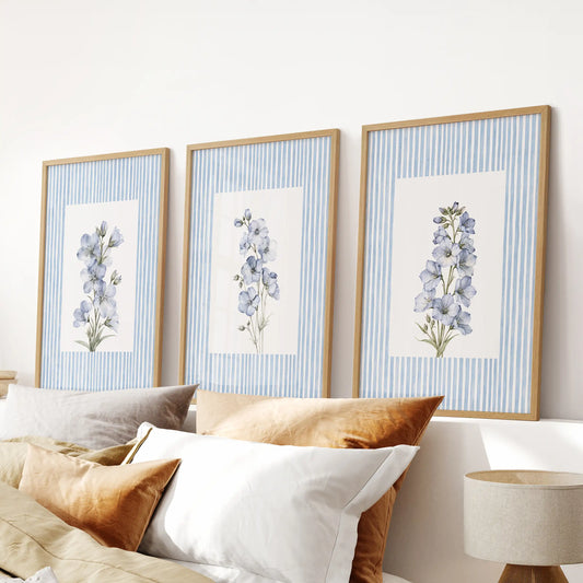 Three-piece pastel botanical wall art set in light oak frames, arranged above the bed in a pale blue bedroom.
