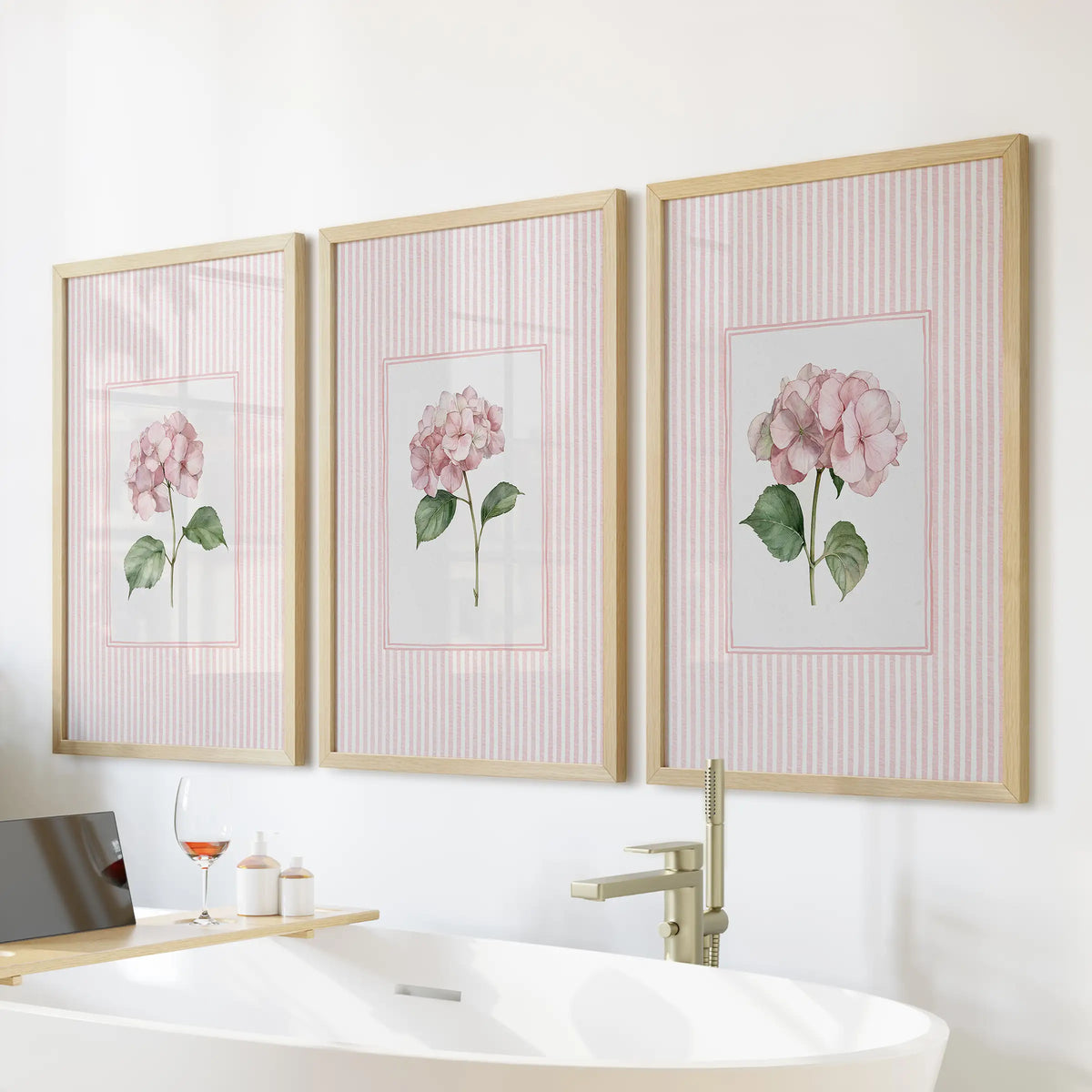 Pink hydrangea botanicals in a three-piece wall art set are displayed in light wood frames over a freestanding tub in a bathroom.