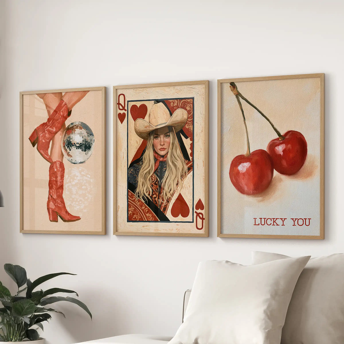 Three framed wall art prints displayed above a sofa in a living room setting. The left print shows red cowboy boots and a disco ball, the center print shows a playing card design with a woman in a cowboy hat, and the right print shows two red cherries with the text 'LUCKY YOU'.