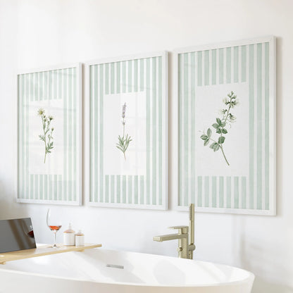 A set of three sage green wildflower wall art prints in white frames hangs over a bathtub in a bathroom.