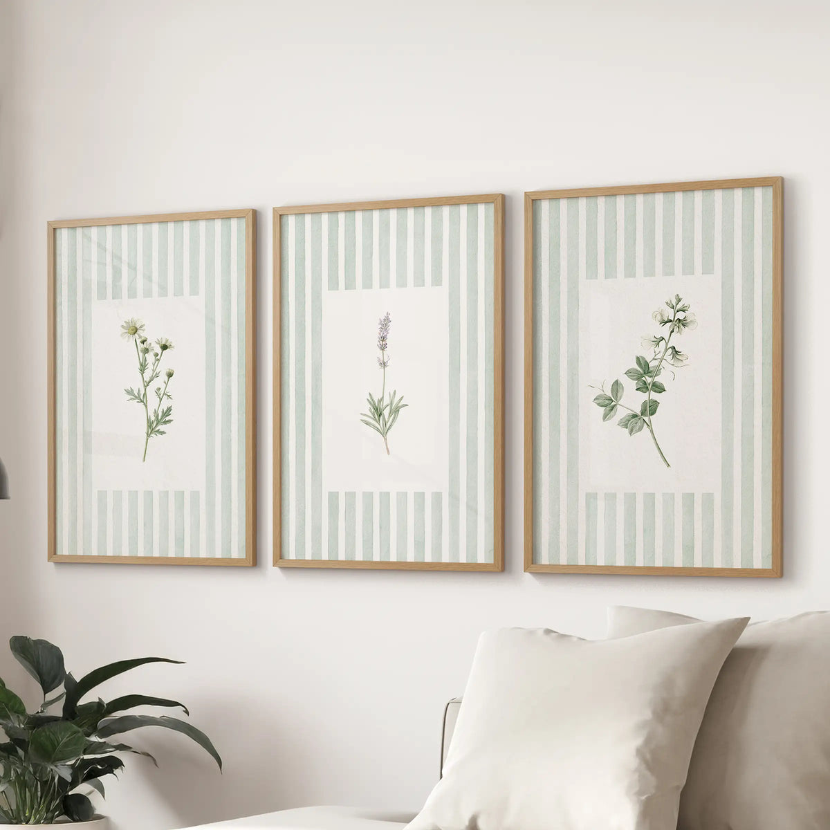 A three-poster set of sage green wildflower wall art in oak frames hangs above a sofa in a family room.