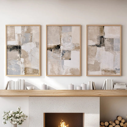 Three framed abstract canvas prints in a light wood frame are displayed side-by-side on a white wall above a fireplace mantel decorated with books, candles, and a small vase of flowers.