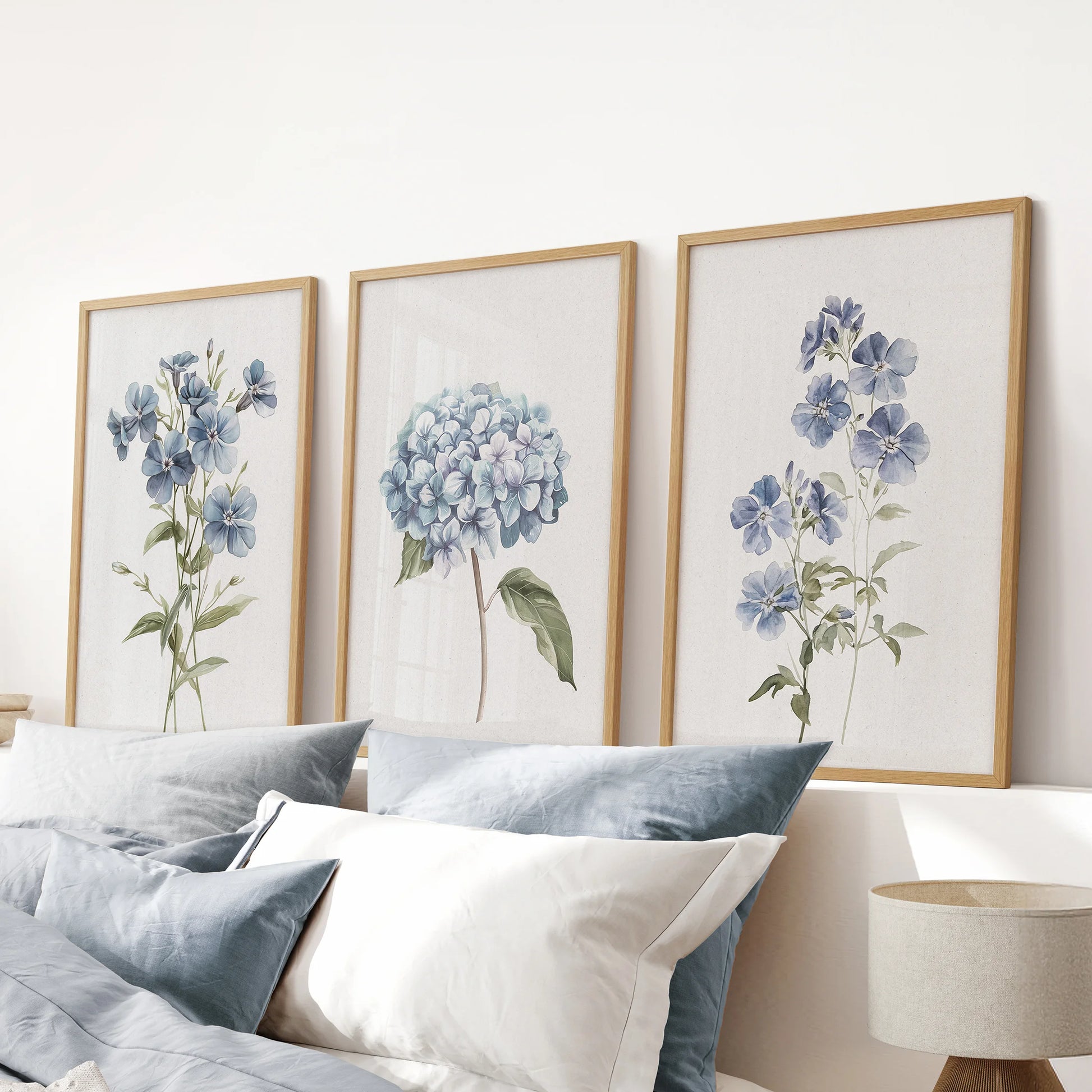 Watercolor botanical florals, a trio of wood-framed wall prints, settle above the headboard in a blue bedroom for a serene, nature-forward accent.