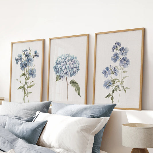 Watercolor botanical florals, a trio of wood-framed wall prints, settle above the headboard in a blue bedroom for a serene, nature-forward accent.