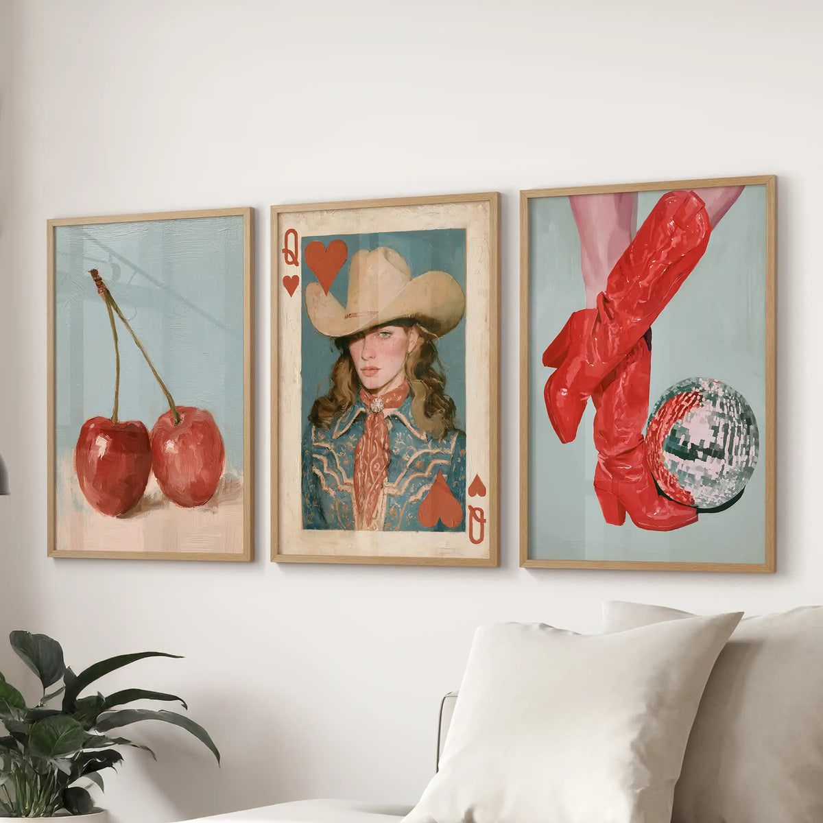 Three framed art prints displayed on a white wall above a sofa. The left print shows two red cherries on a light blue background, the center print is an Ace of Hearts card featuring a woman in a cowboy hat and western clothing, and the right print shows red cowboy boots and a patterned ball against a pale blue background.